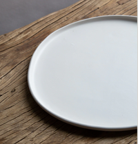 The Urban Dinnerware Matte White plate by Alex Marshall Studios is partially visible on a rustic wooden surface, highlighting the contrast between its handmade ceramic finish and the wood’s natural grain.
