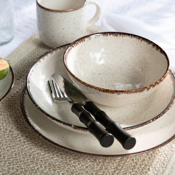 A rustic place setting features a speckled cream bowl, plate, and mug, paired with Blue Pheasant’s Montecito Flatware 5 Piece Setting in Dark Brown, all arranged atop a textured woven placemat.