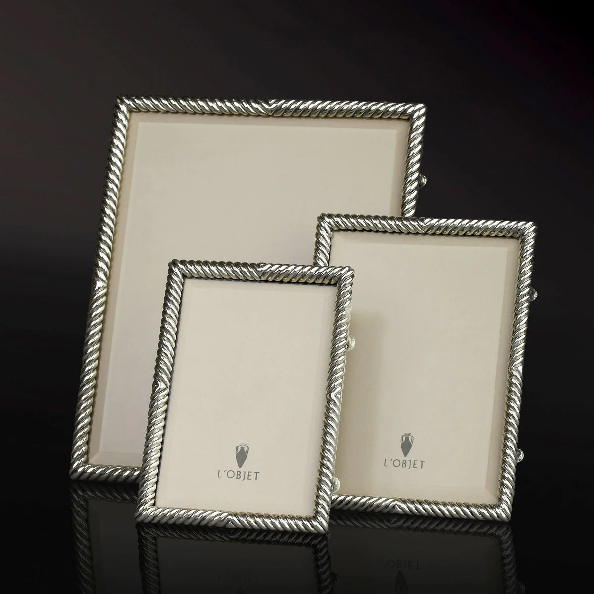 Three empty Deco Twist Platinum Frames by L'Objet, each a different size, stand upright on a glossy black surface. The handcrafted silver frames feature a twisted rope design and beige inserts labeled L'Objet.