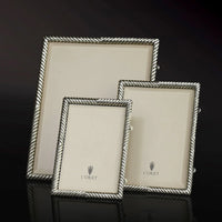 Three empty Deco Twist Platinum Frames by L'Objet, each a different size, stand upright on a glossy black surface. The handcrafted silver frames feature a twisted rope design and beige inserts labeled L'Objet.