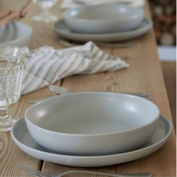 A close-up of a wooden dining table set with Costa Nova Pacifica Dinnerware in Oyster Grey, paired with a striped napkin, silver cutlery, and a glass of water. The elegant dinnerware is beautifully arranged for dining.
