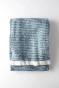 The Four Sided Pinstripe Fringe Throw by A Soft Idea is a folded blue and white wool blend with a subtle houndstooth pattern and white fringe on one end, shown against a plain white background.