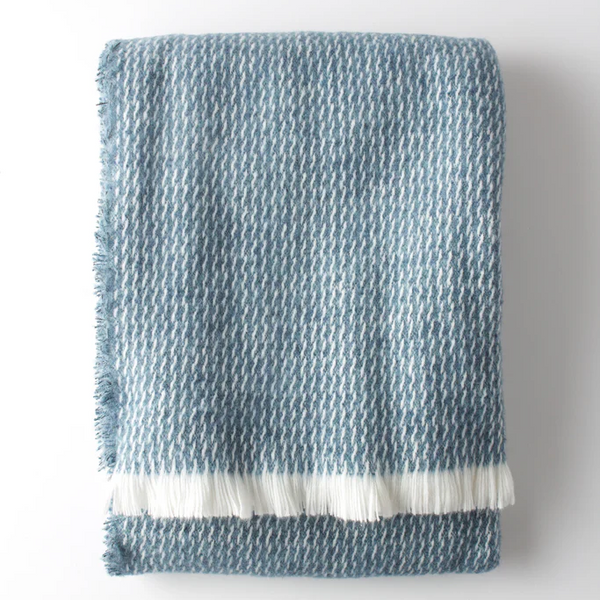 The Four Sided Pinstripe Fringe Throw by A Soft Idea is a folded blue and white wool blend with a subtle houndstooth pattern and white fringe on one end, shown against a plain white background.