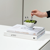 A hand selects a green olive with an Anna New York Striations Silver Cocktail Pick from a decorative bowl resting atop the books Paradises of Colombia and AD at 100 on a white surface.