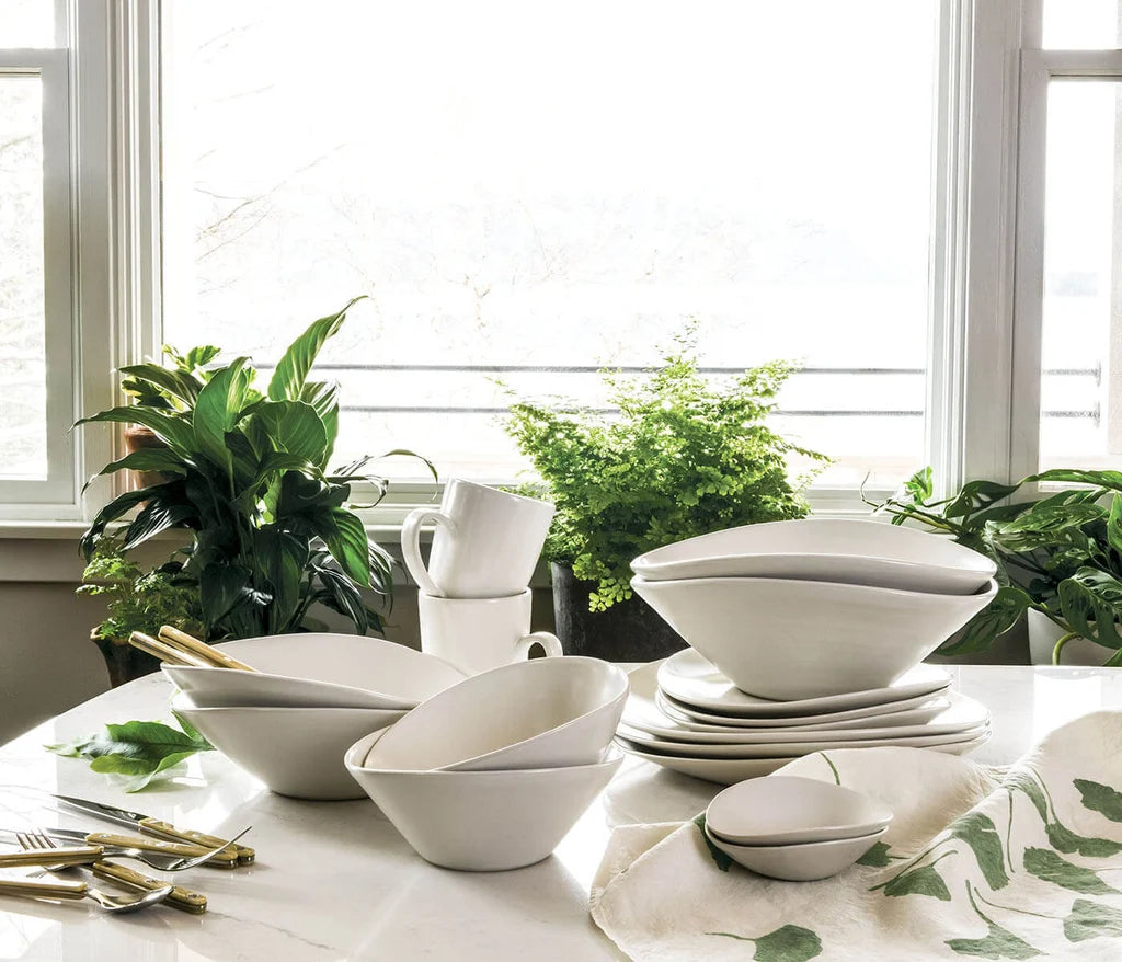 The Simon Pearce Barre Dinnerware Alabaster set, made in the USA, showcases sleek white dishes and bowls artfully arranged on a marble countertop near a window, accented by green leafy plants and gold utensils.