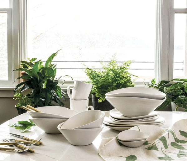 The Simon Pearce Barre Dinnerware Alabaster set, made in the USA, showcases sleek white dishes and bowls artfully arranged on a marble countertop near a window, accented by green leafy plants and gold utensils.