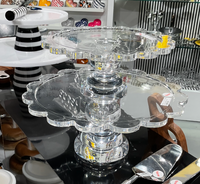 Two Inspired Generations Coco Scalloped Cake Servers, crafted from durable resin acrylic and featuring elegant scalloped edges, are stacked on a display table in a store filled with various kitchenware items such as plates, utensils, and bowls visible in the background.