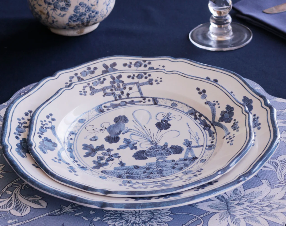 The Beatriz Ball Faenza Melamine Dinnerware Blue & White - Set of 4 features stacked plates with intricate blue and white patterns, styled with a matching placemat, clear wine glass, and blue-patterned bowl.