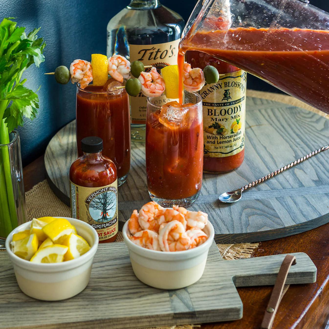 A pitcher pours Bloody Mary mix into a glass among vodka, hot sauce, garnished drinks, shrimp, lemon wedges, and celery—all artfully set on the JK Adams Ash Driftwood Round Serving Board.