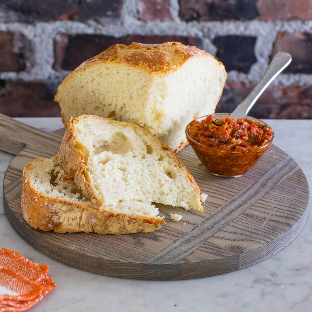 A rustic loaf of bread, partially sliced, rests on a JK Adams Ash Driftwood Round Serving Board with a small glass bowl of chunky red tomato spread and a spoon. A brick wall serves as the background.