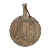 The JK Adams Ash Driftwood Round Serving Board features a handle with a brown leather loop and displays the natural grain of ash wood, highlighted by a stylish driftwood finish.