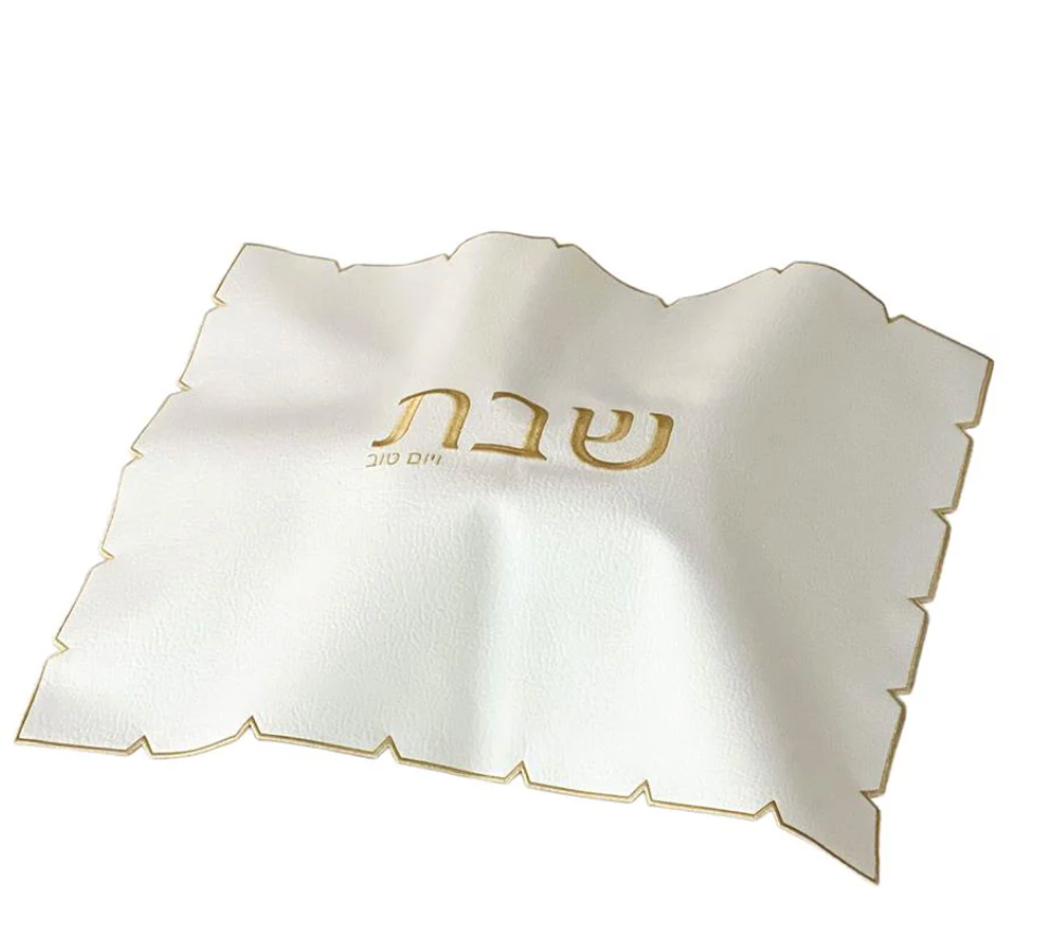 The BT-Shalom White Embroidered Leatherette Challah Cover-Gold features a scalloped, laser-cut edge, gold trim, and "Shabbat" elegantly embroidered in gold at the center.