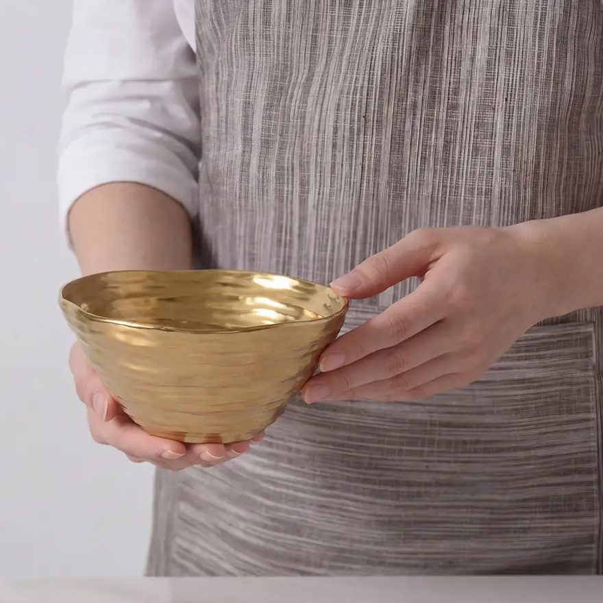 A person in a gray striped apron holds the Dorrato Gold Bowl - by PAMPA BAY, which is matte gold and dishwasher safe, standing behind a white surface.
