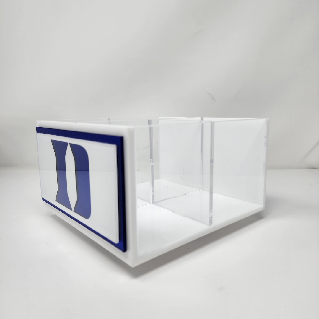 The Eye Candy College Spinner, a clear acrylic desk organizer with three compartments and a bold blue and white D logo on one side, sits on a white surface against a white background.