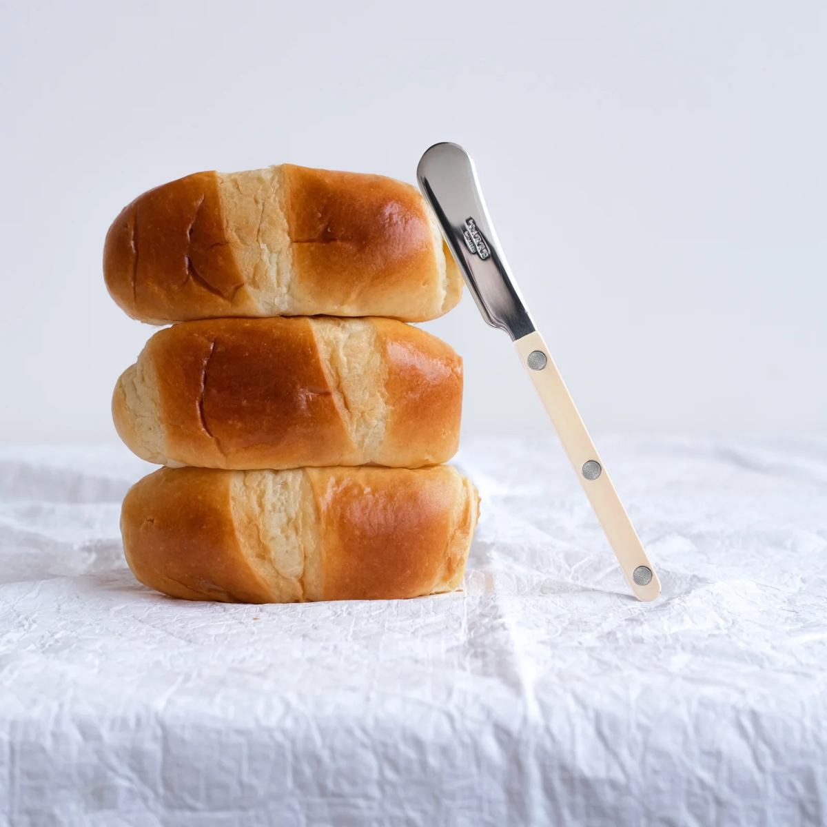 Three stacked bread rolls rest on a white surface, with the Sabre Bistrot Pearly Butter Spreader – Pearl elegantly leaning against them on the right. A soft, light-colored background enhances the simplicity of the scene, making it ideal for highlighting your vibrant Sabre tableware.