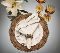 An elegant table setting featuring Kim Seybert’s Swag Napkin Gunmetal & Silver Set of 4, gold-beaded chargers, marble-patterned plates, gold napkin rings, amber glasses, and a white lily on a reflective surface.