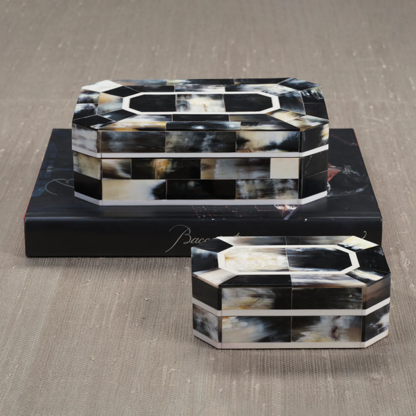 The Zodax Villagio Horn Box Black & Ivory, featuring faceted edges, is stacked on a dark book atop a textured beige surface.
