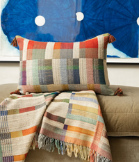 A Threads Soft Goods Lexi Handwoven Pillow 16 x 24 and a matching striped, tasseled throw sit on a beige sofa, set against abstract blue wall art.