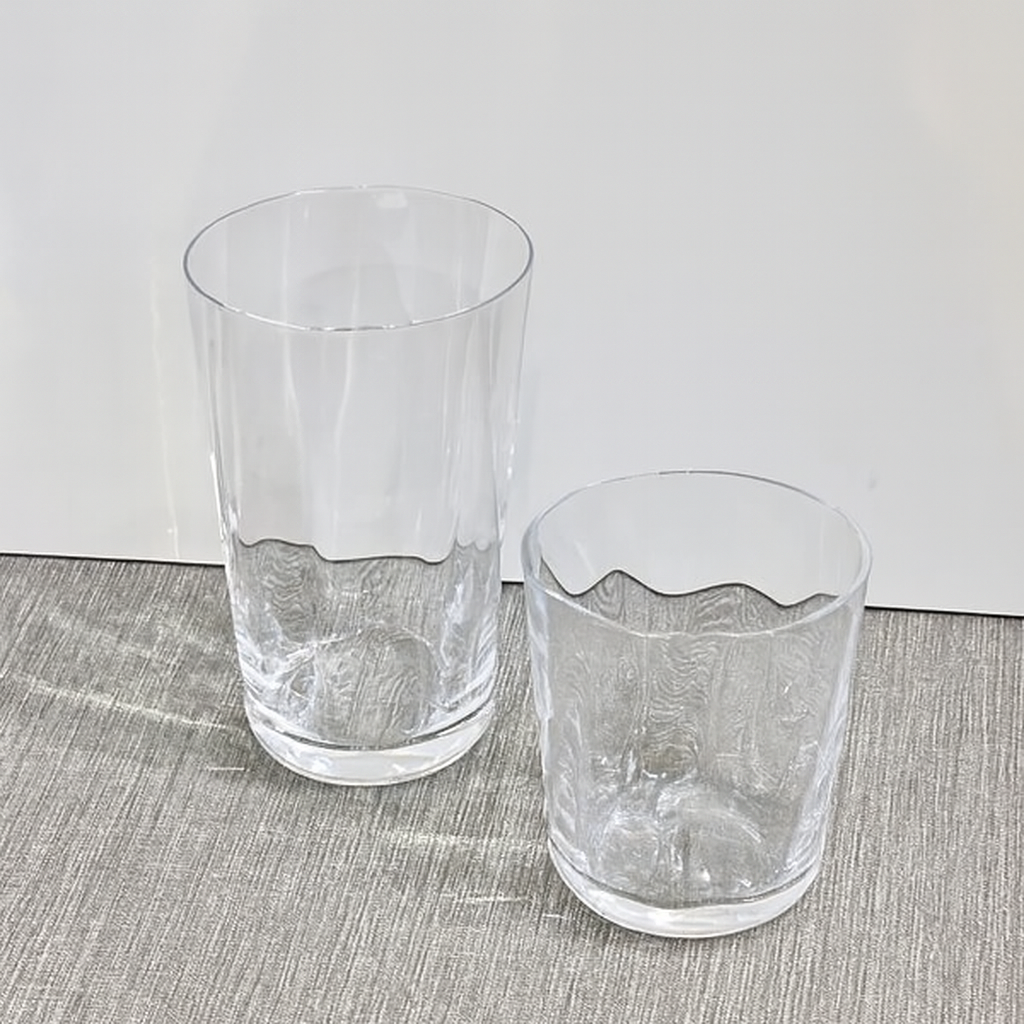 Two empty, clear Optic Everyday Cups by Gary Bodker—one tall, one short—are placed side by side on a light gray textured surface against a plain white background.