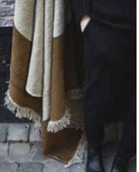 A person dressed in black stands on cobblestone beside the Libeco Home Gus Throw, a brown and beige fringed blanket with natural stripes in beeswax brown, partially covering their legs.