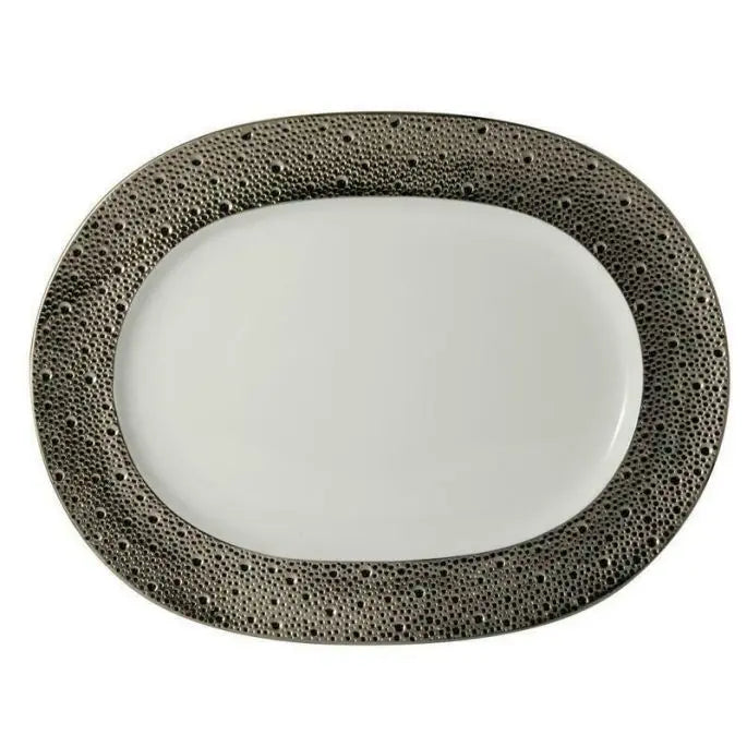The Ecume Platinum Dinnerware by Bernardaud features an oval white plate with a textured, dark metallic border adorned with small, irregular dots—ideal for a contemporary table setting or enhancing your Bernardaud collection.