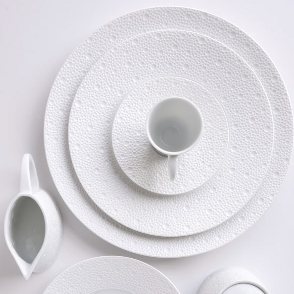 Bernardaud’s Ecume White Dinnerware—dinner plates, teacup, and creamer—features a subtle dotted texture and is elegantly arranged on a white surface for a refined monochromatic table setting.