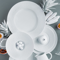Bernardaud's Ecume White Dinnerware—including textured plates, a bowl with white candies, a sleek pitcher, and a white apple—are arranged with painted and cut passion fruits on a gray surface for vibrant contrast.