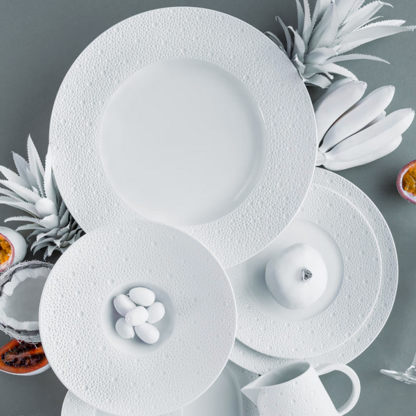 Bernardaud's Ecume White Dinnerware—including textured plates, a bowl with white candies, a sleek pitcher, and a white apple—are arranged with painted and cut passion fruits on a gray surface for vibrant contrast.
