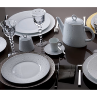 Elegant table setting featuring Bernardaud Ecume White Dinnerware, including textured plates, cups, and teapot, paired with clear patterned wine glasses, silverware, and neutral placemats on a glossy dark surface.