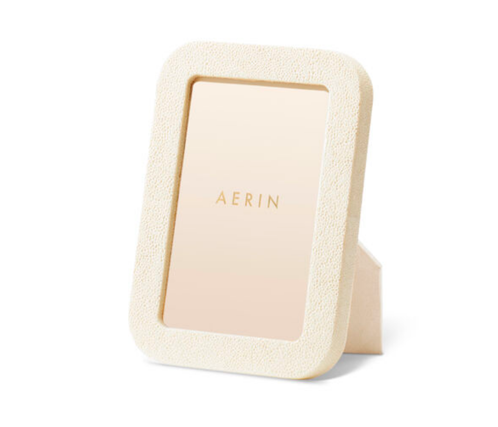 The Modern Shagreen Frame in cream by Aerin is a rectangular, embossed picture frame, standing upright on a white surface and featuring a centered gold "AERIN" placeholder card.