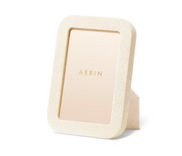 The Modern Shagreen Frame in cream by Aerin is a rectangular, embossed picture frame, standing upright on a white surface and featuring a centered gold "AERIN" placeholder card.