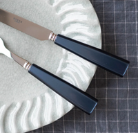 A close-up of the Sabre Icone Flatware 5 Piece Setting with dark, rectangular handles, resting on a textured pale plate featuring a wavy pattern on a gray checkered tablecloth.