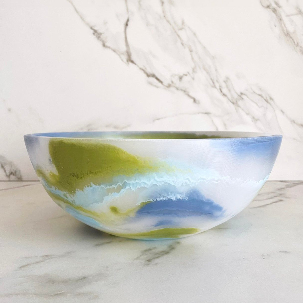 The Redondo Bowl Portofino Blue by CDMX / PAOLA VALLE has a smooth ceramic finish with abstract swirls of blue, green, and white, displayed on a white marble surface with a marbled background.