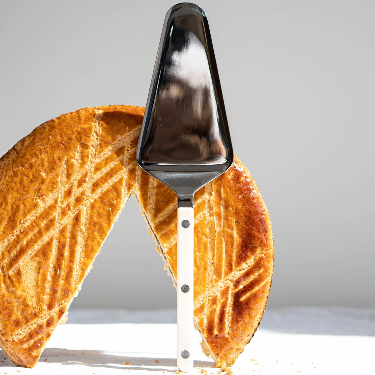 A Sabre Bistrot Solid Tart Server rests in a golden-brown pie with a slice removed, revealing a diamond pattern on top and bringing vintage flair to the presentation.