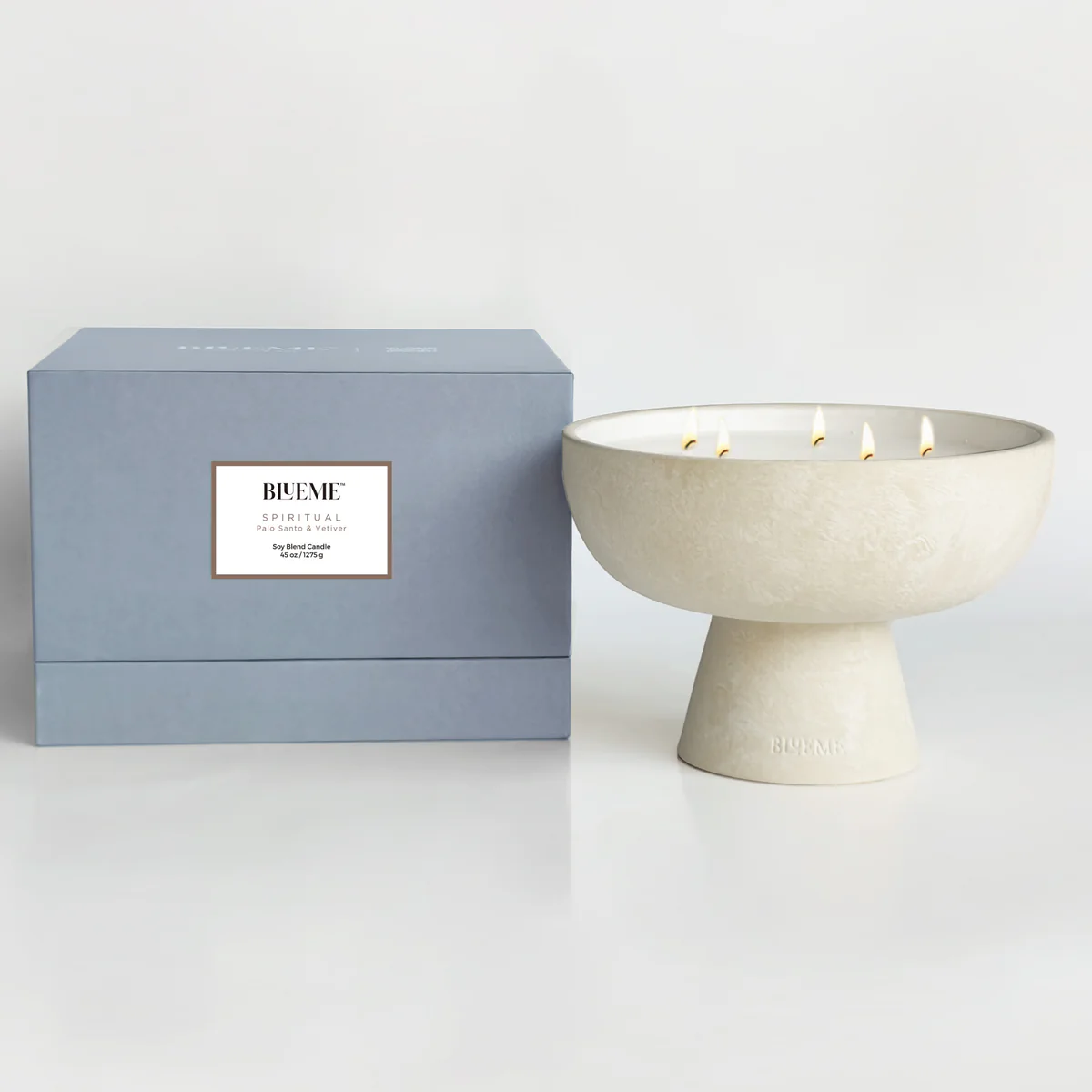 A large, off-white ceramic Spiritual Candle by Blueme, made with a soy wax blend and five wicks, sits next to a light blue BUEME SPIRITUAL box, evoking calming wellness on a simple, light background.