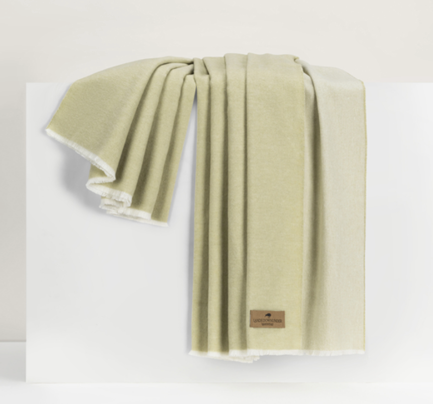 A light green Lands Downunder Aspen Reversible Throw with white fringed edges drapes elegantly over a white surface. A brown leather logo tag is sewn onto one corner, adding a stylish touch to this soft, cozy blanket.