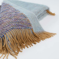 The Coco Throw by Mantas Ezcaray is a soft, woven scarf with blue and purple patterned sections and tan fringed ends, crafted from luxurious fabric and displayed on a white background.