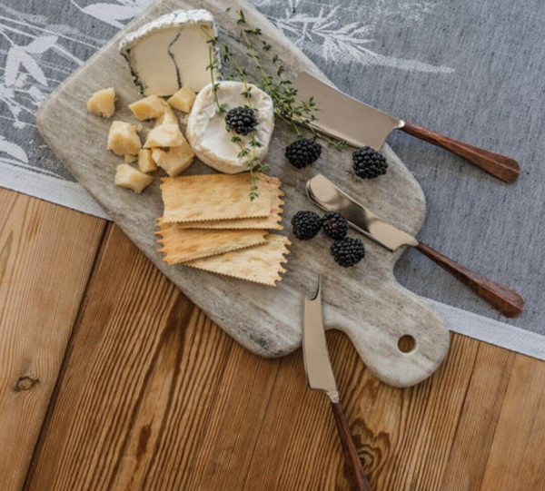 The Simon Pearce Marble Board Medium displays cheese wedges, round crackers, blackberries, and herbs with cheese knives on a wooden table topped with a grey and white patterned tablecloth.