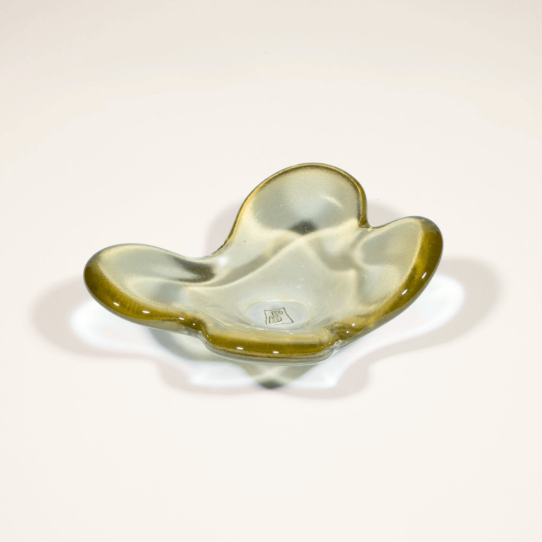 The Cloud Dish by Tyler Barry Design is a hand-blown glass bowl with an organic, wavy form and olive-green tint. It sits on a white surface, casting a soft shadow, and features a small, subtle mark at its center for decoration.