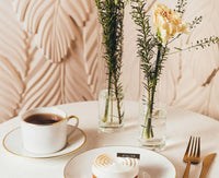 A delicate dessert with torched meringue is served on Bernardaud's Palmyre Dinnerware beside a gold fork, knife, and tea. Two glass vases with greenery and a cream-colored flower lend understated elegance to the refined tablescape.