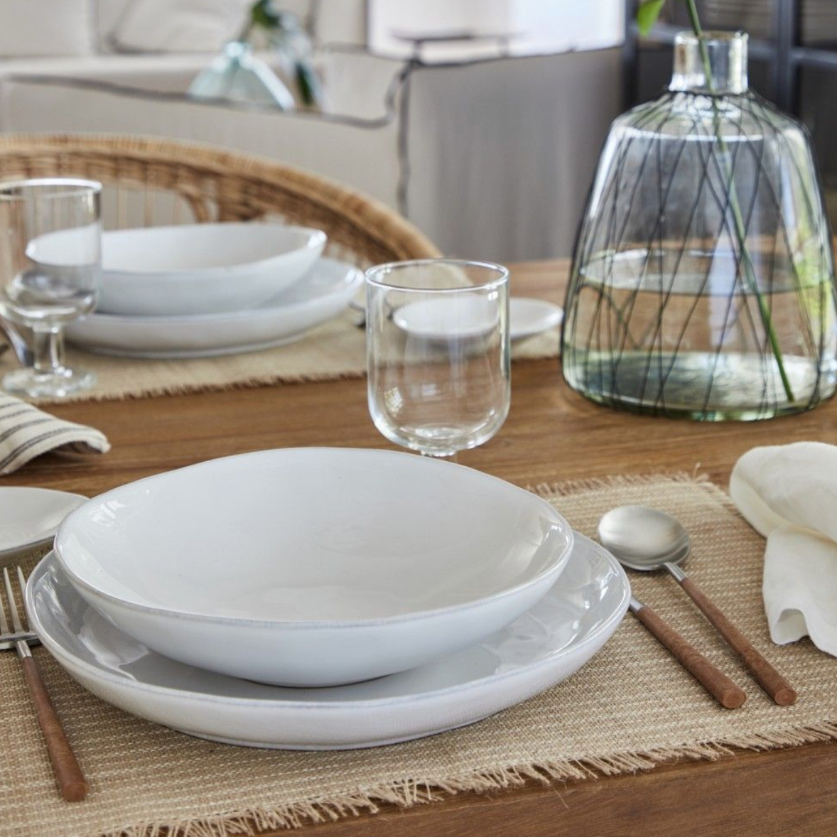 A close-up of a wooden dining table set with modern tableware, featuring Casafina/Costa Nova's Livia Dinnerware White plates, bowls, glasses, silverware with wooden handles, a beige placemat, and a large glass vase with greenery.