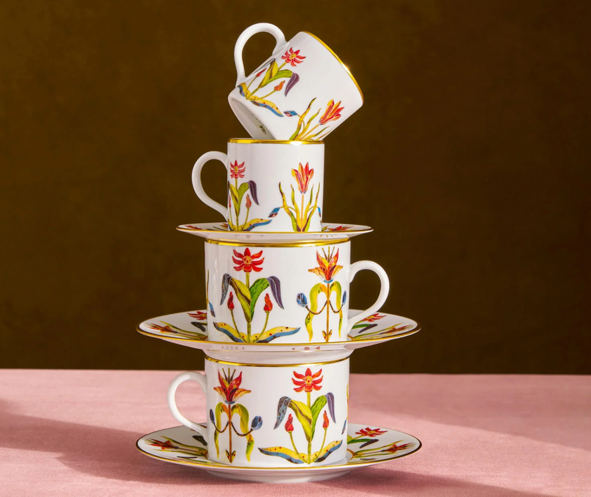 A stack of three handcrafted L’Objet Grand Tour Dinnerware porcelain teacups and saucers from Portugal, decorated with colorful floral patterns and 24k gold accents.