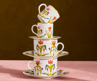 A stack of three handcrafted L’Objet Grand Tour Dinnerware porcelain teacups and saucers from Portugal, decorated with colorful floral patterns and 24k gold accents.