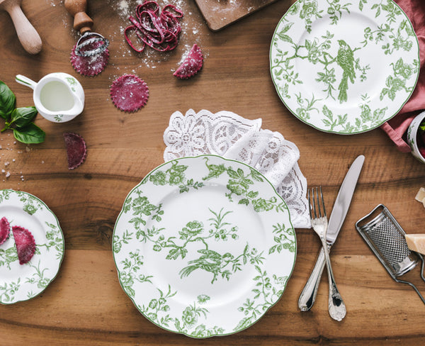 A wooden table set with Bernardaud Albertine Dinnerware: green and white plates, a knife, fork, lace napkin, cheese grater, small jug, purple ravioli, and scattered flour add poetic charm.