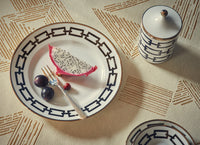 A plate from the Ginori 1735 Catene Nero Dinnerware collection, holding a slice of dragon fruit, two grapes, and a red berry, sits on a patterned tablecloth beside a matching lidded ceramic jar. A fork rests on the plate.