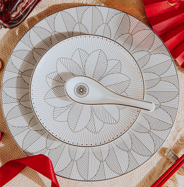 The Christofle Malmaison Imperiale Porcelain Dinnerware Platinum set features a white plate and matching bowl with geometric black leaf patterns, complemented by a coordinated spoon. Red ribbons and chopsticks add elegance to the table setting.