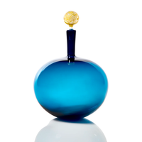 The Cariati Carmella Decanters Steel Blue & Gold features a round, deep blue glass bottle with a tall, narrow neck and decorative yellow stopper, shown against a white background with a reflective surface.