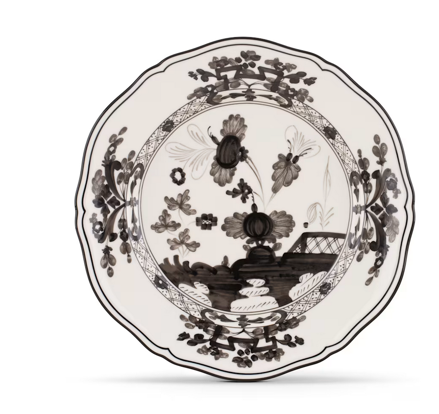 The Oriente Italiano Albus dinnerware by Ginori 1735 is an Italian porcelain plate with a scalloped edge, decorated in black floral patterns and a landscape on white, featuring flowers, leaves, and a building inspired by Ginori Oriente.
