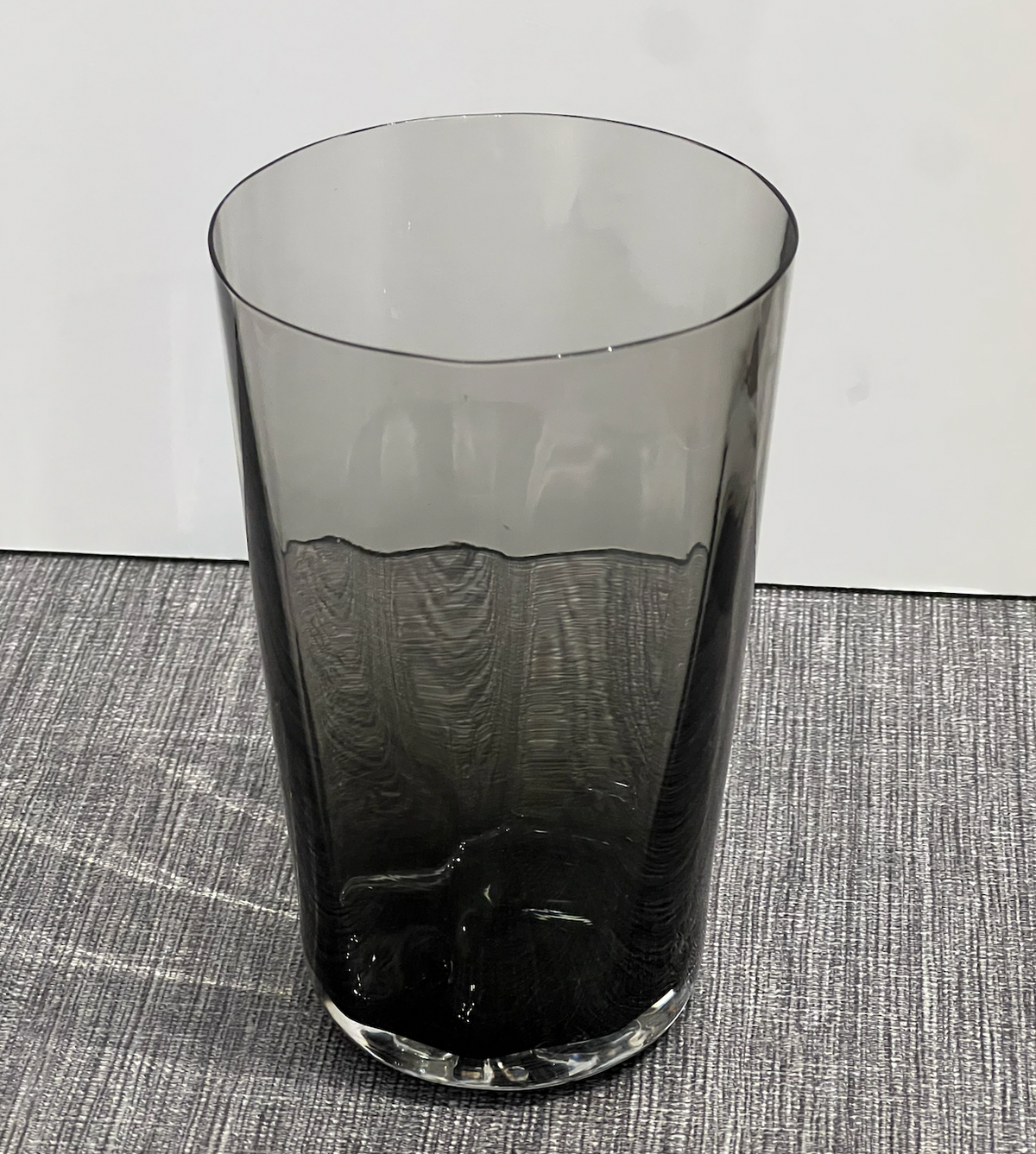 The Optic Cups Charcoal by Gary Bodker, a tall, cylindrical handcrafted glassware in translucent dark gray, sits on a textured gray surface against a plain white background.
