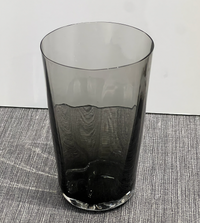 The Optic Cups Charcoal by Gary Bodker, a tall, cylindrical handcrafted glassware in translucent dark gray, sits on a textured gray surface against a plain white background.